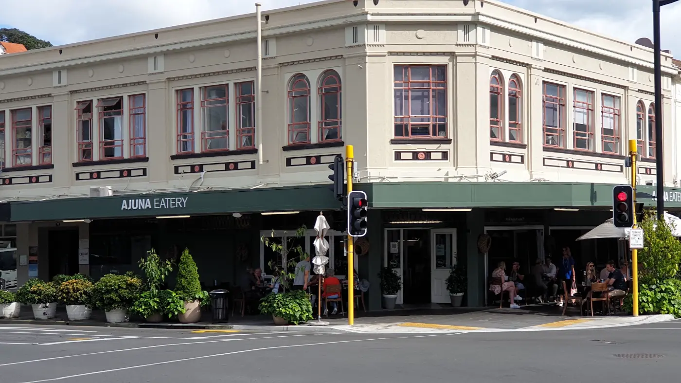 Ajuna Eatery on the corner of a cream two-storey building with arched red-framed windows, a green awning, potted plants and people at tables outside